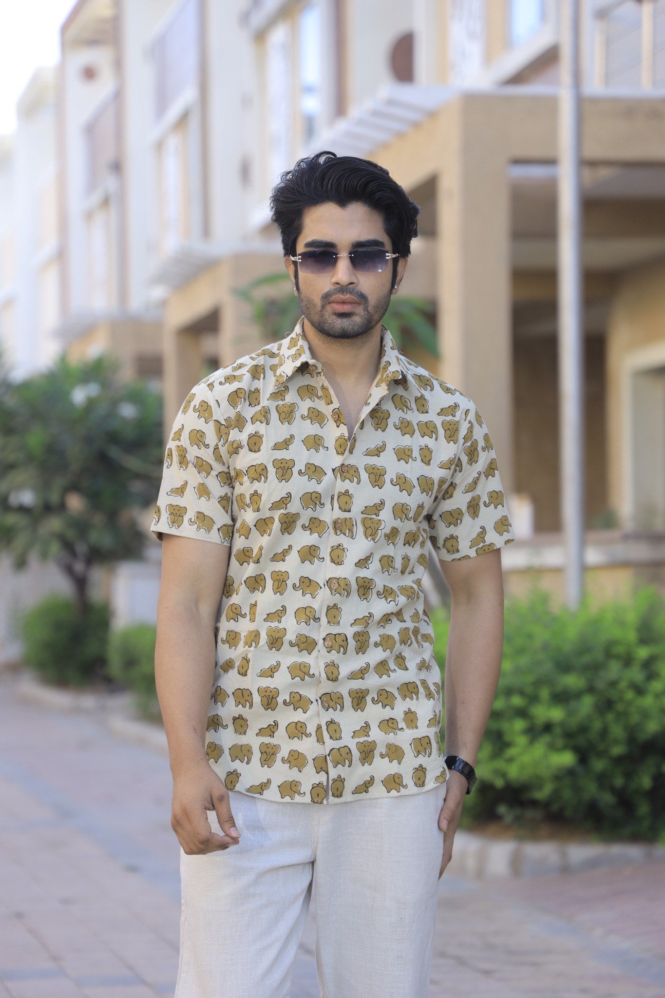 A man wearing a beige short-sleeve button-up shirt with a repeating brown animal print, paired with white trousers and dark sunglasses, standing outdoors on a paved walkway with a blurred residential background.